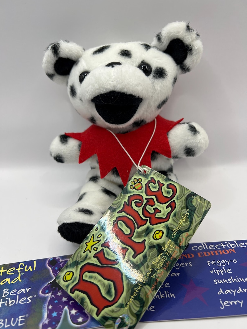 May include: A small, white and black dalmatian-patterned plush bear wearing a red collar. The bear is holding a tag that reads "Grateful Dead Bean Bear collectibles" and "Liquid Blue".