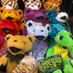 May include: A collection of colorful plush teddy bears and a turtle, all with different patterns and designs. The bears are wearing various outfits, including a blue and white striped shirt, a red and white striped shirt, and a green and white striped shirt. The turtle is wearing a yellow shell with a green and white pattern. The bears and turtle are all sitting on a black surface. The tags on the bears and turtle read "Grateful Dead Bears" and "Liquid Blue".