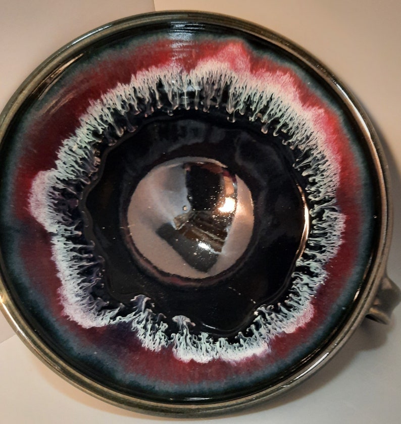 May include: A large, round ceramic bowl with a dark blue glaze and a white and red rim. The bowl has a unique, swirling design in the glaze.