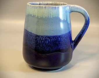 May include: A ceramic mug with a gradient of blue hues. The mug transitions from a light blue at the rim to a deep navy at the base. It features a curved handle and a wide, sturdy base, suitable for hot beverages.