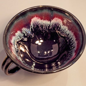 May include: A dark purple ceramic bowl with a white and red glaze. The bowl has a unique, abstract design and a handle on the side.