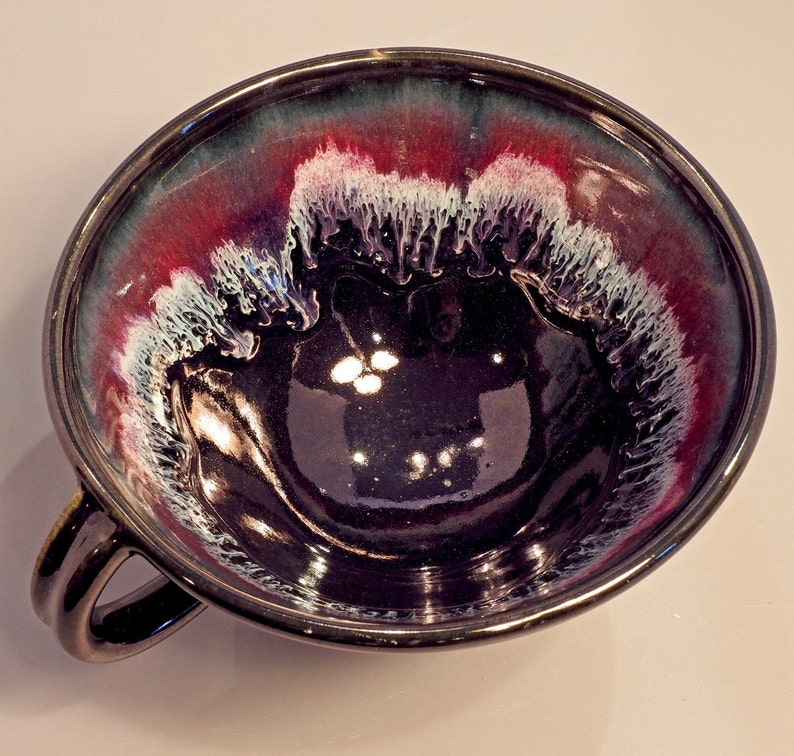 May include: A dark purple ceramic bowl with a white and red glaze. The bowl has a unique, abstract design and a handle on the side.