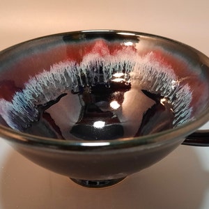 May include: A black ceramic bowl with a red and white glaze. The bowl has a handle and a rounded bottom.