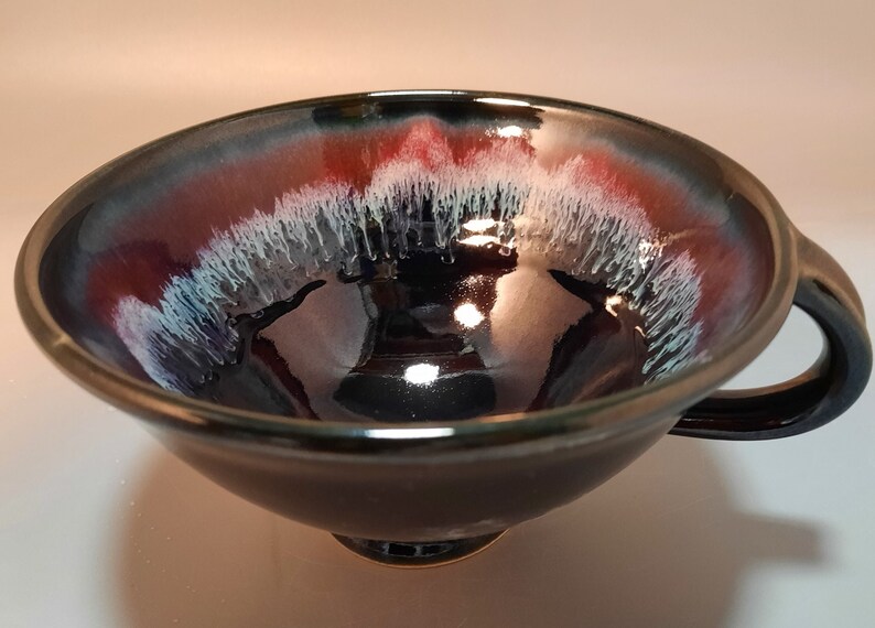 May include: A black ceramic bowl with a red and white glaze. The bowl has a handle and a rounded bottom.