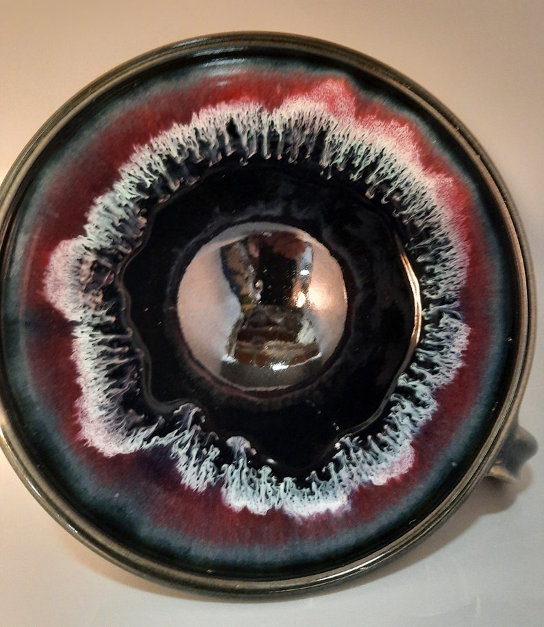 May include: A ceramic mug with a dark blue glaze and a white and red rim. The inside of the mug is glazed with a shiny, reflective finish.