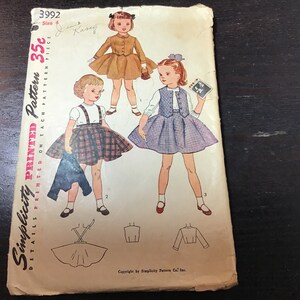 May include: Vintage Simplicity sewing pattern number 3992 for girls' dresses. The pattern includes three different styles: a plaid skirt with suspenders, a purple skirt with a vest, and a brown skirt with a jacket. The pattern is printed on a white background with black text and illustrations.