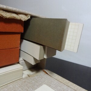 May include: A close-up of a stack of books with orange covers, a stack of white books, and a stack of brown and white books. The books are in a box with a light brown fabric lining.