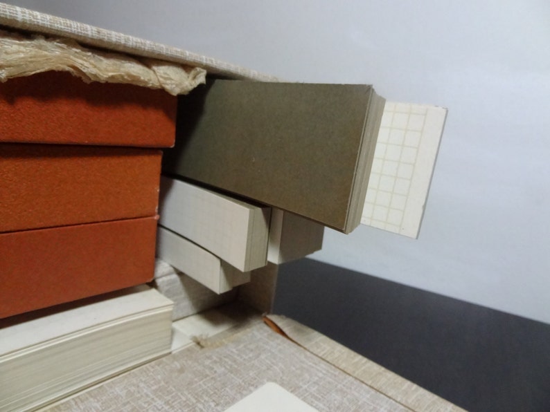 May include: A close-up of a stack of books with orange covers, a stack of white books, and a stack of brown and white books. The books are in a box with a light brown fabric lining.