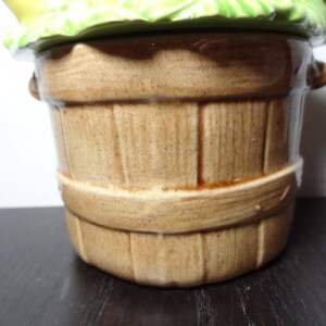 May include: A brown ceramic cookie jar shaped like a wooden bucket with a green leafy lid. The lid has a yellow flower design.