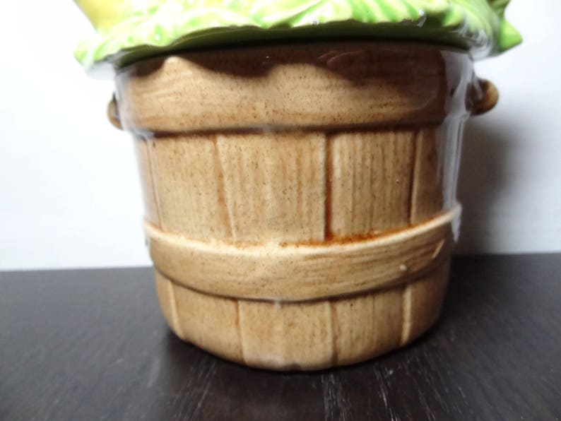 May include: A brown ceramic cookie jar shaped like a wooden bucket with a green leafy lid. The lid has a yellow flower design.