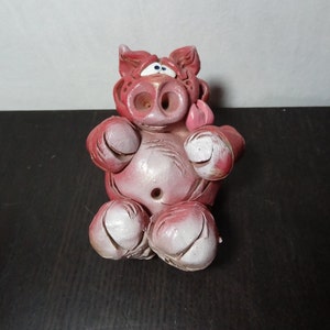 May include: A pink ceramic pig figurine with a white belly and a tongue sticking out. The pig has a hole in its belly.