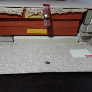 May include: A vintage wooden storage box with a beige fabric exterior and a red leather handle. The box is open and contains several stacks of white paper cards. The box has a label that reads "11/2" Black Letters & Numerals".