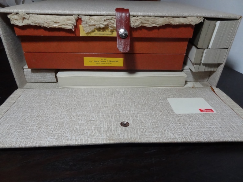 May include: A vintage wooden storage box with a beige fabric exterior and a red leather handle. The box is open and contains several stacks of white paper cards. The box has a label that reads "11/2" Black Letters & Numerals".