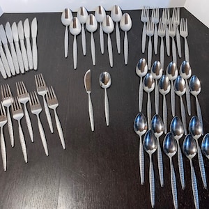 May include: A collection of stainless steel flatware, including knives, forks, and spoons, arranged on a dark surface. The utensils have textured handles and a simple, elegant design, suitable for a formal dining setting.