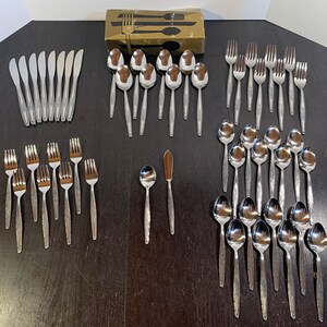 May include: A set of silverware in a silver finish, including forks, spoons, and knives. The silverware is arranged in rows on a dark wooden surface.