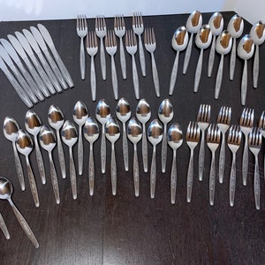 May include: A collection of stainless steel silverware, including spoons, forks, and knives, arranged on a dark surface. The silverware has a floral pattern on the handles. The set includes butter knives and a variety of spoons and forks.