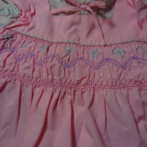 May include: A pink dress with a white and pink plaid trim. The dress has a smocked bodice with a purple and white embroidered design.