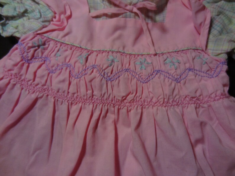 May include: A pink dress with a white and pink plaid trim. The dress has a smocked bodice with a purple and white embroidered design.