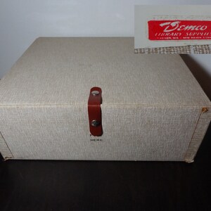 May include: A beige cardboard box with a brown leather handle and a label that reads "Demco Library Supplies" in red letters. The box has a textured surface and a "Push Here" label on the front.