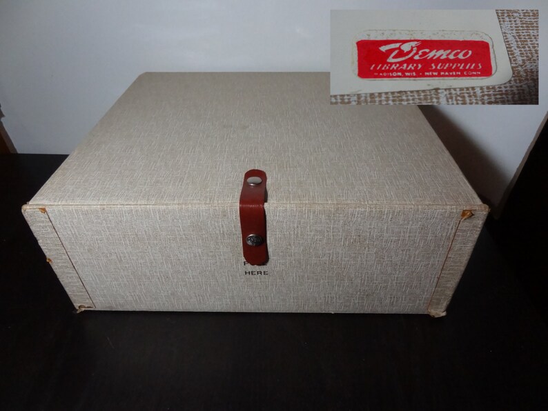 May include: A beige cardboard box with a brown leather handle and a label that reads "Demco Library Supplies" in red letters. The box has a textured surface and a "Push Here" label on the front.