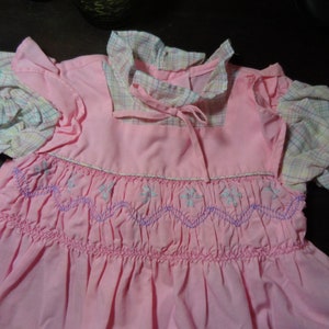 May include: A pink smocked dress with a plaid collar and sleeves. The dress has a white and purple embroidered design.