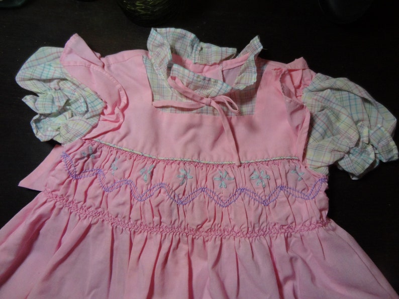 May include: A pink smocked dress with a plaid collar and sleeves. The dress has a white and purple embroidered design.