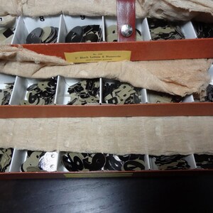 May include: Three wooden boxes with compartments filled with black letter and number stencils. The boxes are stacked on top of each other. The top box has a label that reads "2" Black Letters & Numerals.  The boxes are made of wood and have a brown finish.
