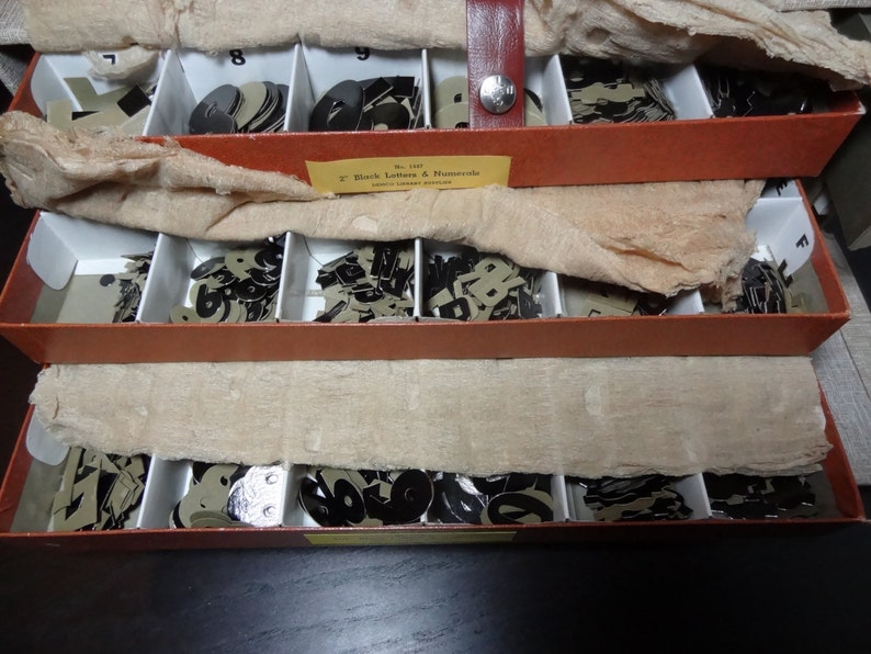 May include: Three wooden boxes with compartments filled with black letter and number stencils. The boxes are stacked on top of each other. The top box has a label that reads "2" Black Letters & Numerals.  The boxes are made of wood and have a brown finish.