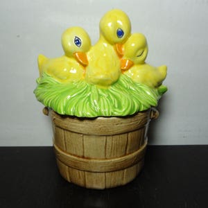 May include: A ceramic cookie jar shaped like a wooden bucket with three yellow ducklings nestled in green grass on top.