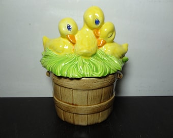 May include: A ceramic cookie jar shaped like a wooden bucket with three yellow ducklings nestled in green grass on top.