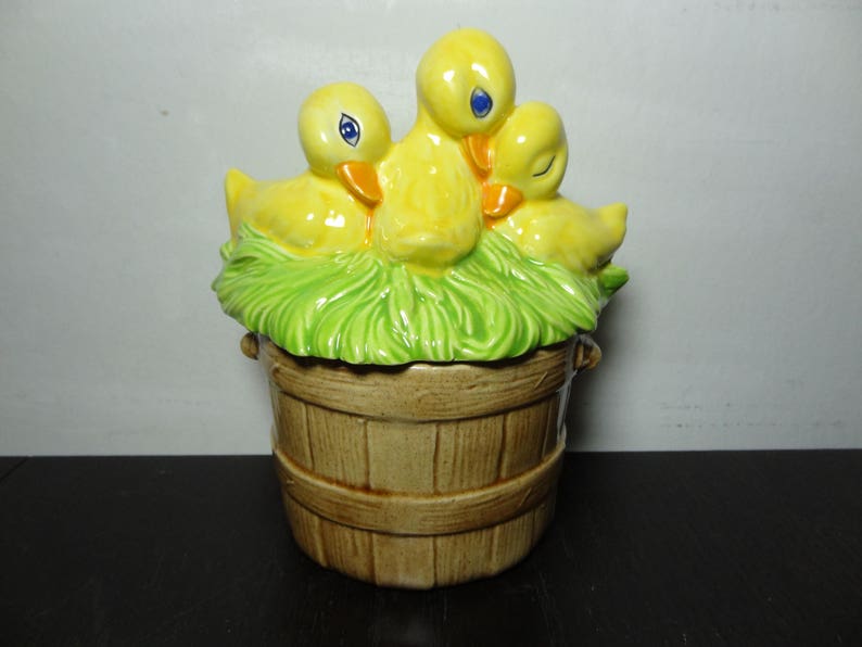 May include: A ceramic cookie jar shaped like a wooden bucket with three yellow ducklings nestled in green grass on top.