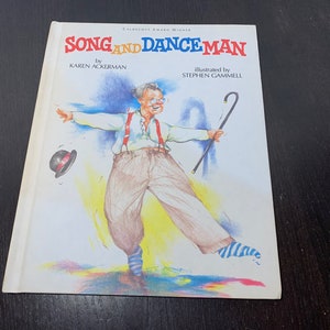 May include: A book cover featuring a colourful illustration of a man in braces and a hat dancing with a walking stick. The title of the book is "Song and Dance Man" by Karen Ackerman, illustrated by Stephen Gammell.