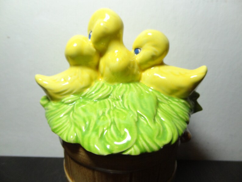 May include: A ceramic birdhouse with three yellow birds perched on a green nest. The birdhouse is shaped like a barrel and has a brown base.