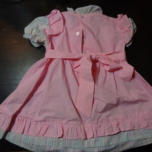 May include: A pink dress with a plaid trim and a large bow in the back. The dress has a ruffled hem and is made of a soft, lightweight fabric.