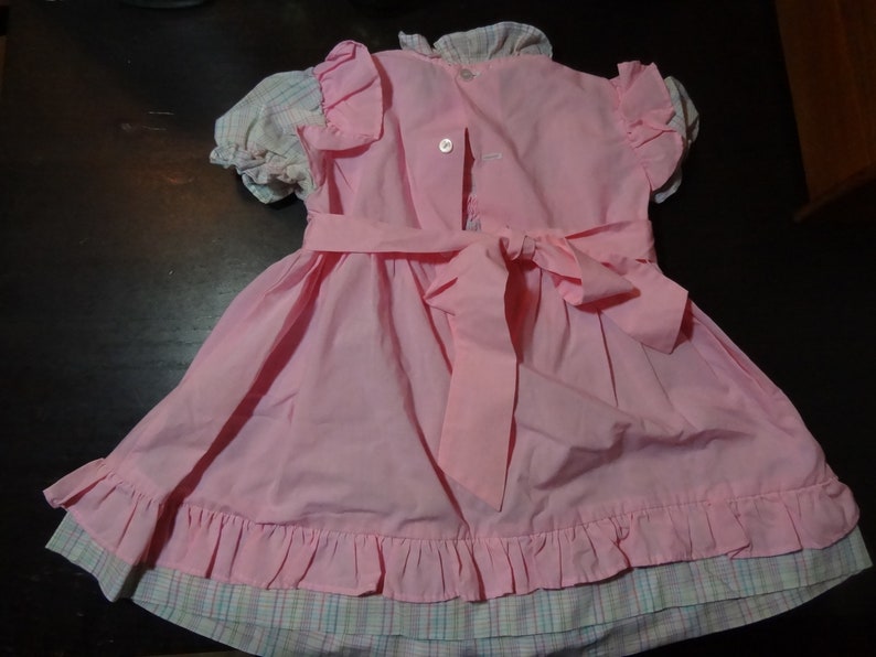May include: A pink dress with a plaid trim and a large bow in the back. The dress has a ruffled hem and is made of a soft, lightweight fabric.