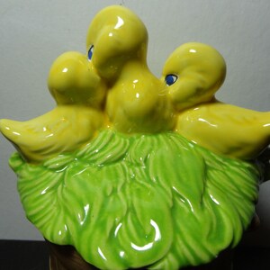 May include: A ceramic figurine of three yellow chicks nestled in a green leafy nest. The chicks have blue eyes and are arranged in a cluster.