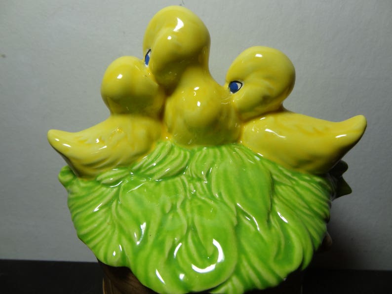 May include: A ceramic figurine of three yellow chicks nestled in a green leafy nest. The chicks have blue eyes and are arranged in a cluster.