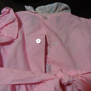 May include: A pink dress with a plaid collar and ruffled sleeves. The dress has a button closure in the back and a sash tie at the waist.