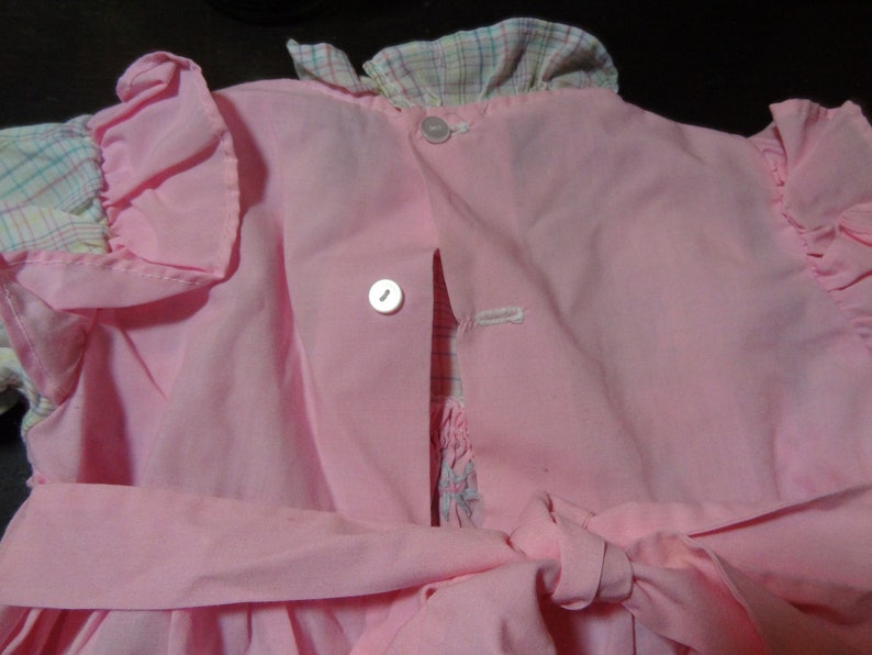 May include: A pink dress with a plaid collar and ruffled sleeves. The dress has a button closure in the back and a sash tie at the waist.