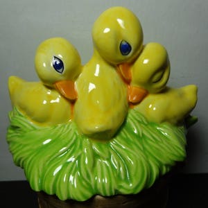 May include: A ceramic figurine of three yellow ducklings nestled in a green nest. The ducklings have black eyes and orange beaks.
