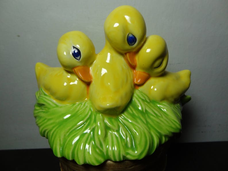 May include: A ceramic figurine of three yellow ducklings nestled in a green nest. The ducklings have black eyes and orange beaks.