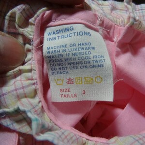 May include: Pink and white plaid fabric with a white tag that says "Washing Instructions" and includes instructions for machine or hand washing, pressing, and bleaching. The tag also includes a size label that says "Taille 3".