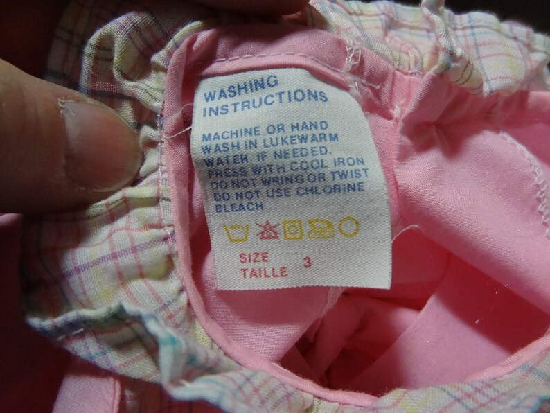 May include: Pink and white plaid fabric with a white tag that says "Washing Instructions" and includes instructions for machine or hand washing, pressing, and bleaching. The tag also includes a size label that says "Taille 3".