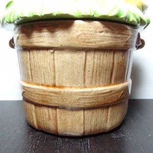 May include: A ceramic cookie jar shaped like a brown wooden basket with a green leafy lid. The lid has a yellow and white floral design.