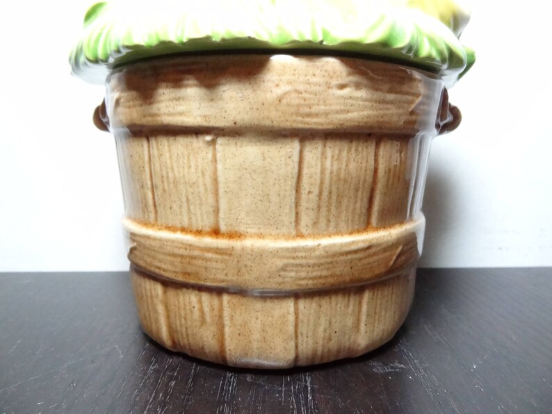 May include: A ceramic cookie jar shaped like a brown wooden basket with a green leafy lid. The lid has a yellow and white floral design.