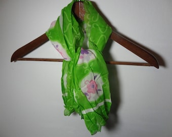 May include: A green silk scarf with white and pink floral designs. The scarf is hanging on a wooden hanger.