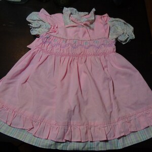 May include: A pink smocked dress with a white and pink plaid trim. The dress has a ruffled hem and a white collar with a bow.