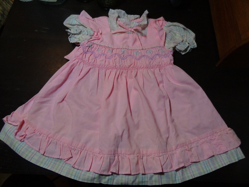 May include: A pink smocked dress with a white and pink plaid trim. The dress has a ruffled hem and a white collar with a bow.