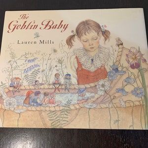 May include: The cover of a children's book titled "The Goblin Baby" by Lauren Mills. The illustration shows a young girl looking down at a group of small goblins in a tub of water. The goblins are wearing clothes and have wings. The girl is wearing a red dress and a white lace collar.