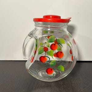 May include: A clear glass pitcher with a red lid and a cherry design. The pitcher has a handle and is decorated with red cherries and green leaves.